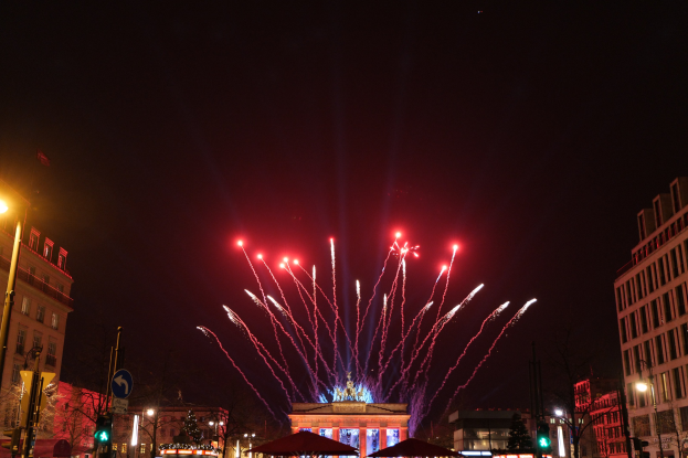 Eine nächtliche Stadtstraße mit dem beleuchteten Brandenburger Tor in Berlin, Deutschland, mit Feuerwerk am Himmel, umgeben von Gebäuden, Bäumen, Laternen, Ampeln, Schildern, Zelten und Menschen.