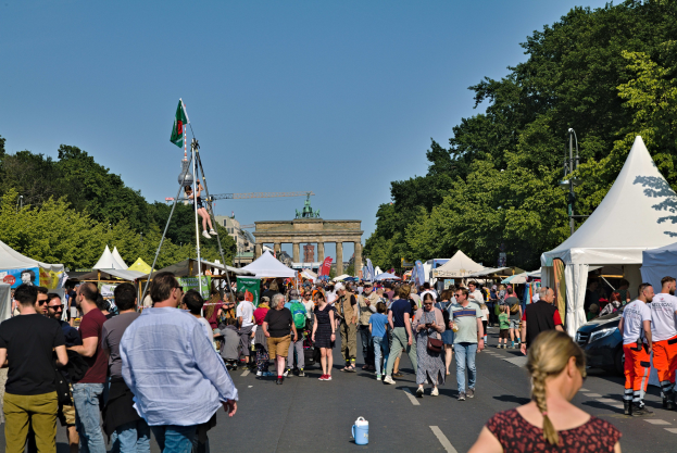 Eine Menschenmenge, die eine von Zelten, Fahrzeugen und Bäumen gesäumte Straße entlanggeht, mit einem Bogen und einem klaren blauen Himmel im Hintergrund und Fahnenmasten auf der linken Seite.