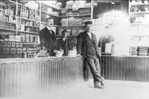 Ein Schwarz-Weiß-Foto von drei Männern, die an einem Tresen in einem Geschäft stehen, mit verschiedenen Gegenständen auf dem Tresen und einem Regal voller Bücher und anderer Gegenstände im Hintergrund während eines Raubs.