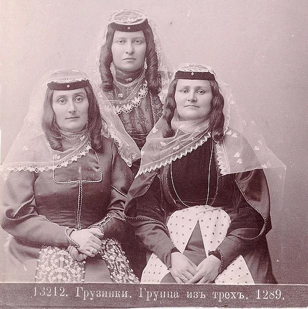 Schwarz-weißes Foto von drei Frauen in traditionellen russischen Hochzeitskleidern mit farbenfrohen Stickereien und Perlen, die vor einer weißen Wand stehen.