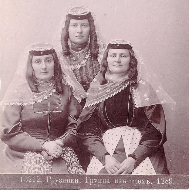 Schwarz-weißes Foto von drei Frauen in traditionellen russischen Hochzeitskleidern mit farbenfrohen Stickereien und Perlen, die vor einer weißen Wand stehen.