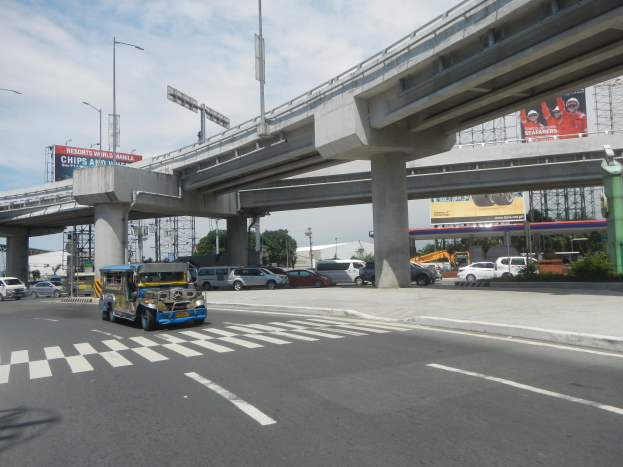 Vielbefahrene Hauptstraße mit Fahrzeugen, Zebrastreifen, Überführung mit Säulen, Laternen, Informationsschildern, Bäumen, Gebäuden und bewölktem Himmel.