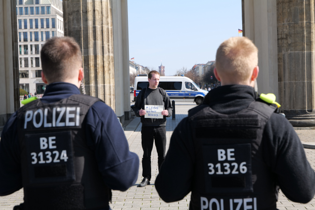Drei Polizeibeamte stehen vor einem Gebäude, ein Mann in der Mitte hält ein Papier, flankiert von Säulen und umgeben von Fahrzeugen, Menschen, Bäumen, Gebäuden und dem Himmel.