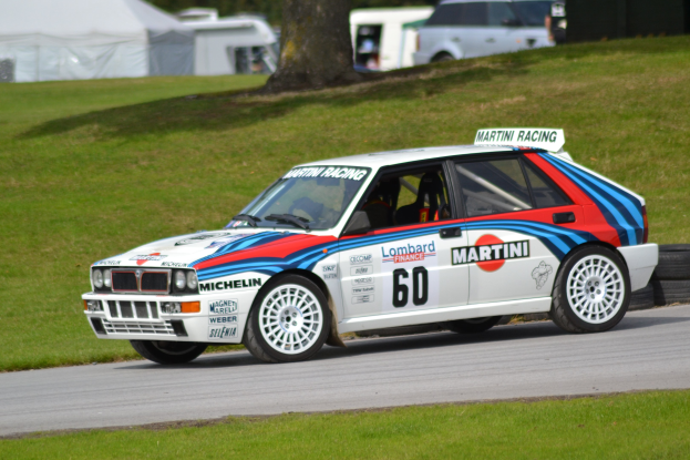 Ein Lancia Delta HF Integrale Evoluzione Group B Rally Auto auf einer Straße mit Gras, Bäumen, Zelten und anderen Fahrzeugen, mit Text und Zahlen auf seiner Karosserie und einem Reifen im Vordergrund.