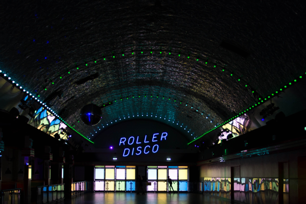 Ein Roller-Disco mit beleuchtetem "Roller-Disco"-Schild, Menschen auf dem Boden, Glastüren im Hintergrund und Säulen links und rechts.