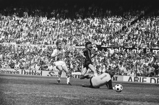 Schwarzes und weißes Foto von Männern, die auf einem Feld Fußball spielen, mit Zuschauern im Hintergrund und einer 'Bundesliga 1978-79 Ajax 1-1 Hannover 96' -Fahne.