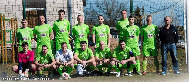 Gruppe von Männern in grünen T-Shirts auf einem Fübballfeld mit einem Ball und einem Tor, umgeben von Bäumen, Gebäuden und Fahrzeugen.