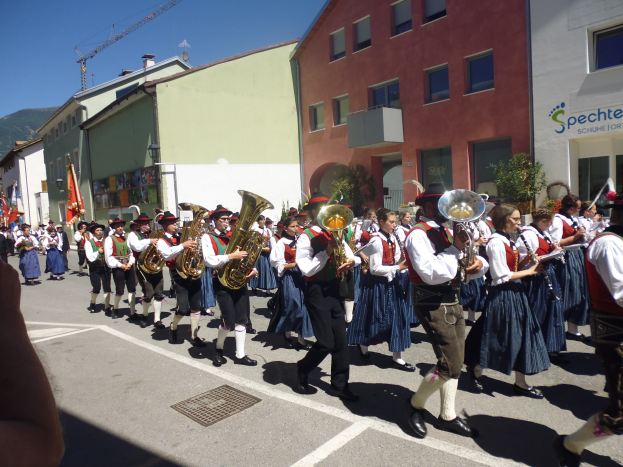 Eine Gruppe von Menschen in traditioneller bayrischer Tracht, die auf einer Straße mit Gebäuden spielen, einige halten Fahnen, mit einem Hügel und einem blauen Himmel im Hintergrund.