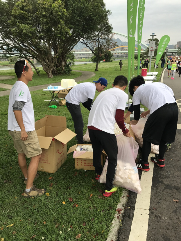 Gruppe von Menschen an einer Straße mit verstreuten Kartons und Flaschen, die an einem Spendenlauf teilnehmen, mit Bäumen, Gebäuden, Polen, Schildern und einem klaren blauen Himmel im Hintergrund.