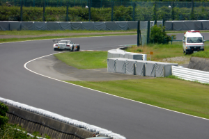 Ein Rennauto fährt eine Rennstrecke entlang mit einem Polizeiwagen im Hintergrund, umgeben von Gras, Pflanzen, Bäumen und Absperrungen, mit einem Fahrzeug auf der rechten Seite und Text unten.