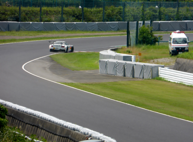 Ein Rennauto fährt eine Rennstrecke entlang mit einem Polizeiwagen im Hintergrund, umgeben von Gras, Pflanzen, Bäumen und Absperrungen, mit einem Fahrzeug auf der rechten Seite und Text unten.