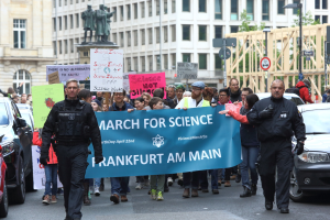 Eine Gruppe von Menschen marschiert mit einer 'March for Science Frankfurt am Main'-Fahne die Straße entlang, mit Autos, Gebäuden, Statuen, Laternen, Schildern und Bäumen im Hintergrund.