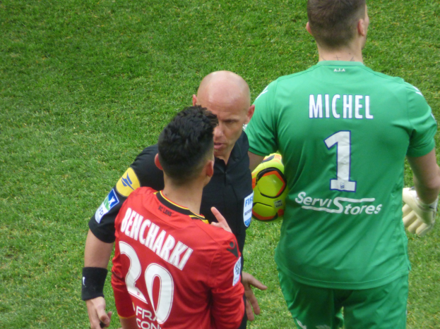Schiedsrichter spricht mit zwei Fußballspielern auf einem saftig grünen Feld, einer hält einen Fußball.