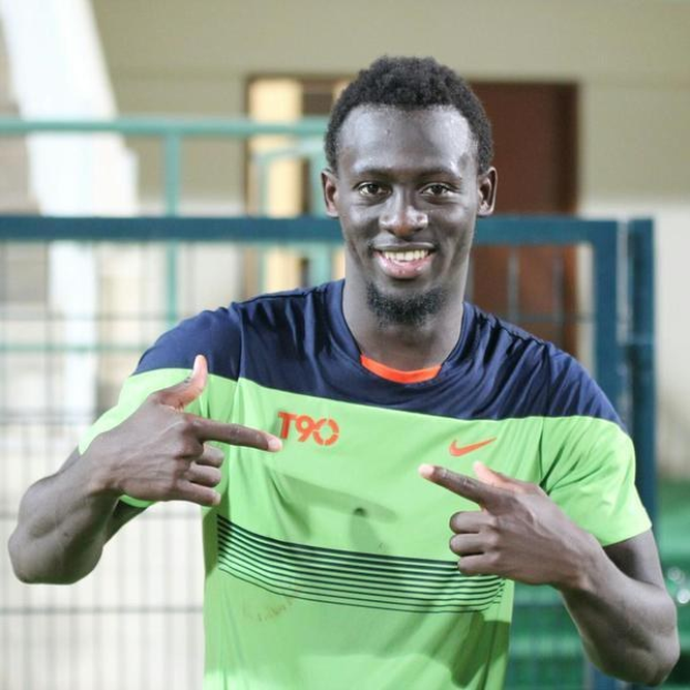 Ein Mann in einem grün-blauen Hemd lächelt und zeigt auf die Kamera, im Hintergrund ist eine leicht unscharfe Reling, Wand und Tür zu sehen.