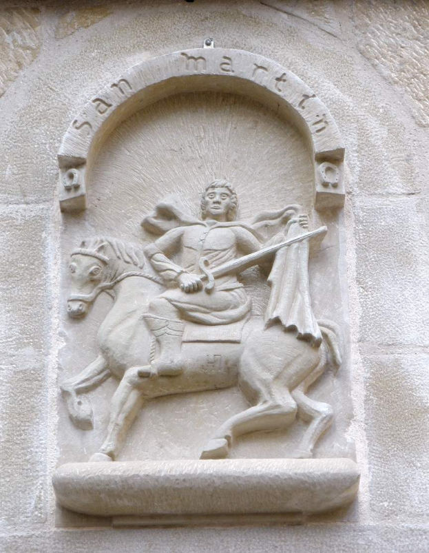 Steinrelief an einer Gebäudewand, das einen Mann auf einem Pferd, wahrscheinlich Sankt Martin, mit einem Schwert in der Hand zeigt, mit Inschrift darunter.