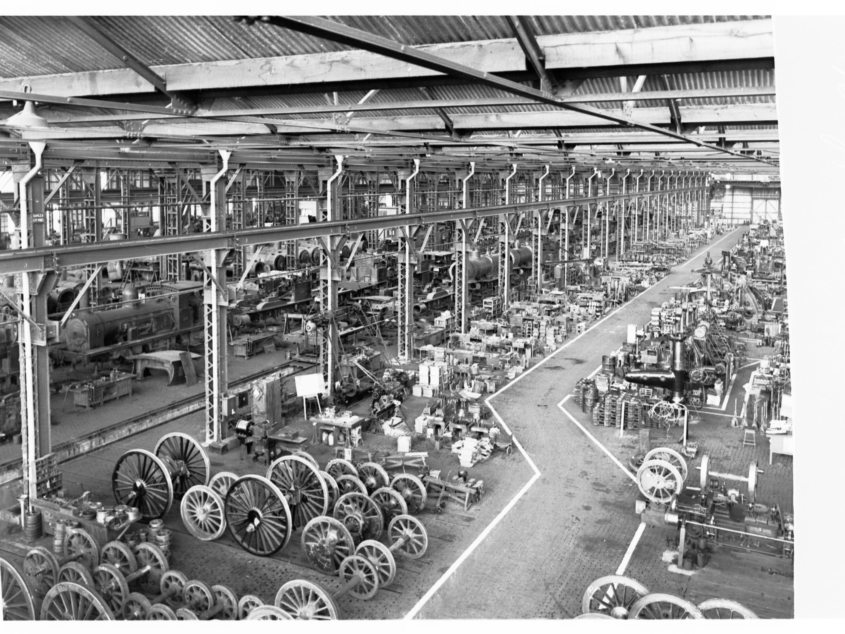 Schwarz-weiß-Foto einer industriellen Fabrikhalle mit Maschinen, einschließlich Rädern, Polen und anderem Equipment, gekrönt von einer Halle.