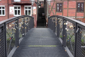 Eine Brücke, geschmückt mit Love Locks, spannt sich vor einem Backsteingebäude, das von Laternen beleuchtet wird, mit Stufen, die zum Gebäude hinaufführen, und Text am unteren Bildrand.