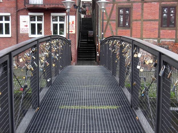 Eine Brücke, geschmückt mit Love Locks, spannt sich vor einem Backsteingebäude, das von Laternen beleuchtet wird, mit Stufen, die zum Gebäude hinaufführen, und Text am unteren Bildrand.