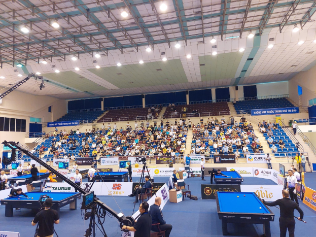 Ein großer Raum mit mehreren Billardtischen, umgeben von sitzenden Spielern, Fenstern an der linken Wand, Deckenbeleuchtung und einer Kameraaufstellung, die auf ein professionelles Billardturnier hinweist.