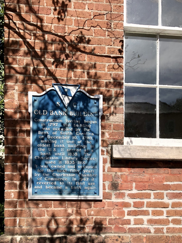 Altes Bankgebäude mit einer blauen Tafel an der Seite, ein Fenster rechts und einen Baum links.
