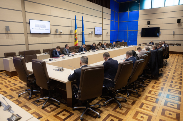 Große Gruppe von Menschen um einen langen Tisch versammelt mit Mikrofonen und Papieren in einer förmlichen Besprechungssituation, mit EU-Flaggen, Fernsehern und Deckenleuchten im Hintergrund.