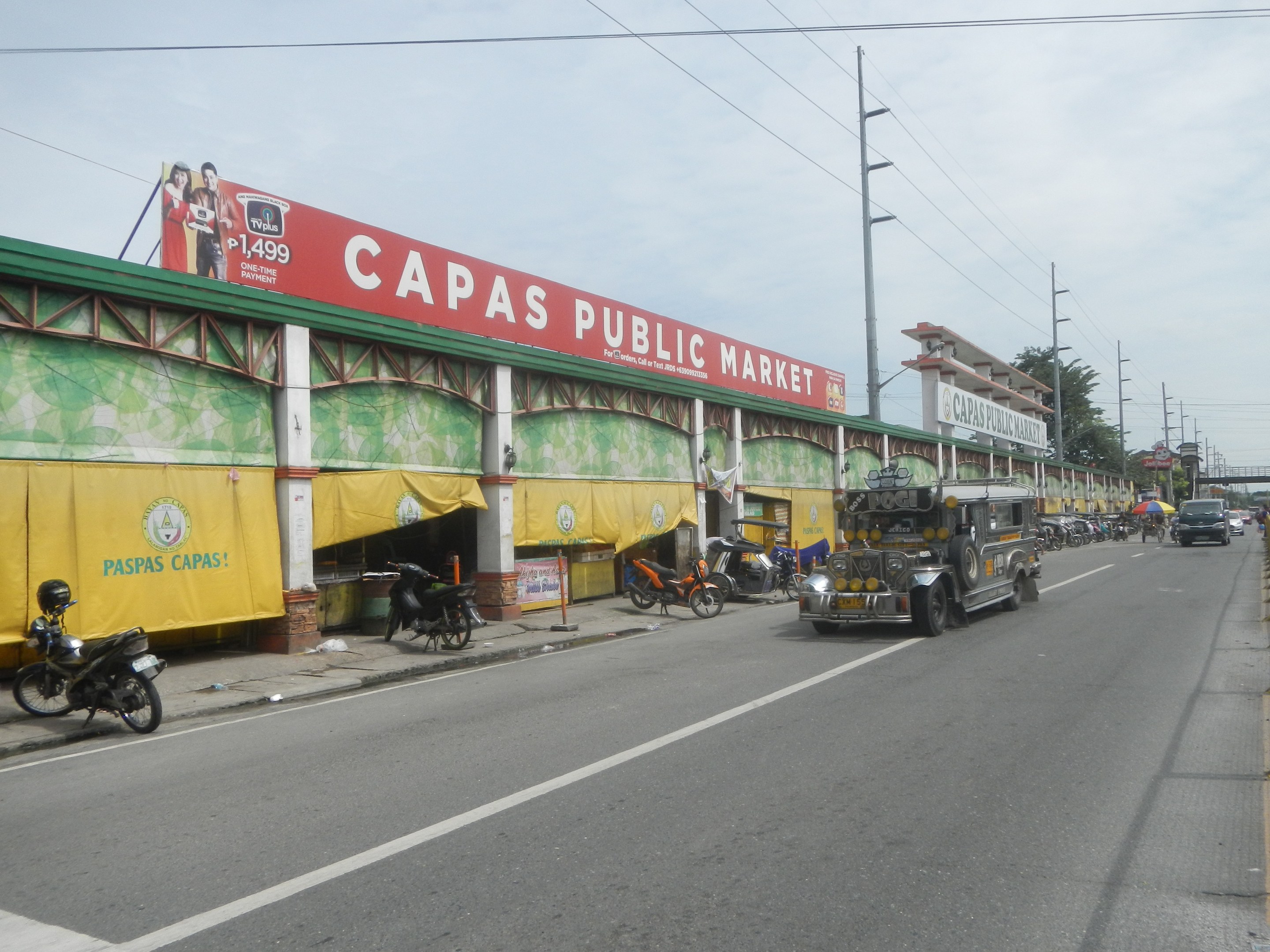 Eine belebte Straße mit Fahrzeugen, einem Gehweg, Strommasten, Gebäuden, Bäumen und einem bewölktem Himmel, mit einem Gebäude mit der Aufschrift "Capas Public Market" im Vordergrund.