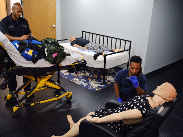 Ein Raum zur Ersten-Hilfe-Ausbildung mit einer Puppe auf einer Trage, zwei Personen auf Betten, einer Person stehend mit einem Gegenstand in der Hand und einer weiteren Person sitzend auf einem Stuhl; eine Tasche auf der Trage und eine Matte auf dem Boden, mit einer Wand und einer Tür im Hintergrund.