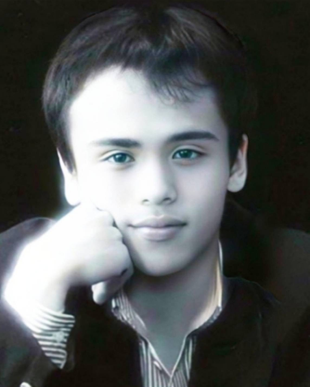 Ein junger Mann in einer schwarzen Jacke posiert selbstbewusst vor einem dunklen Hintergrund, mit einem entschlossenen Gesichtsausdruck und leuchtenden Augen.