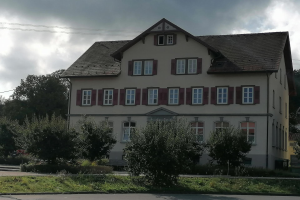 Großes weißes Haus mit roten Fensterläden, umgeben von Bäumen, Pflanzen und Gras, mit einer Straße davor und Stromleitungen im Hintergrund, als Gästehaus zum Verkauf angeboten in Baden-Württemberg.