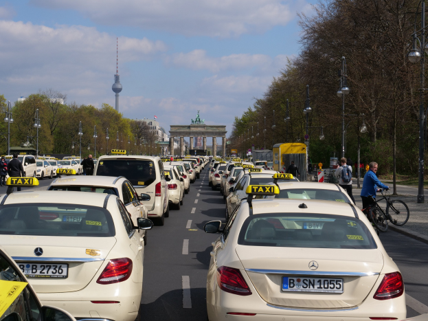 Eine lange Reihe von Taxis, die an einer belebten Straße in Berlin, Deutschland, geparkt sind, mit Fahrradfahrern und Füßgängern auf dem Gehweg, flankiert von Bäumen und Laternen, und Gebäuden, einem Bogen und einem Turm im Hintergrund unter einem bewölktem Himmel.
