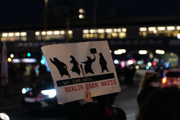 Eine Person hält ein Schild mit der Aufschrift "Berlin gegen Nazis" vor einer Menge und Fahrzeugen auf einer Straße, während Gebäude das Bild erhellen.