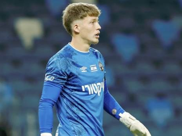 Ein junger Torwart in einem blauen Trikot steht auf einem Fußballfeld und trägt ein blaues T-Shirt, weiße Handschuhe und scheint während eines Spiels im Einsatz zu sein.