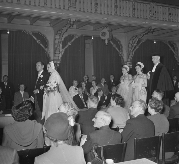 Schwarz-weiß-Foto einer Hochzeitszeremonie in einem großen Raum mit sitzenden und stehenden Gästen, einige halten Blumensträuße, Tische mit Tassen und Gegenständen sowie Vorhänge und Deckenleuchten im Hintergrund.