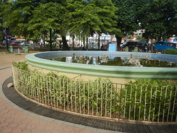 Ein parkümfangener Park mit einem zentralen Brunnen, umgeben von B├Ąumen, Pflanzen, einem Schild, Fahrzeugen, Passanten, Flaggenmasten, Geb├Ąuden mit Fenstern und einem bew├Âlktem Himmel.