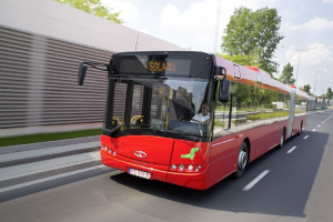 Ein roter Bus fährt auf einer Straße neben einem Gebäude, in dem sich eine Person befindet, während sich auf der linken Seite eine Wand befindet und im Hintergrund Bäume, Laternenpfähle und ein bewölkter Himmel befinden.