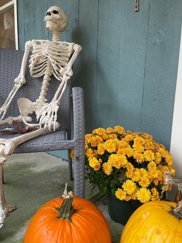 Ein Skelett sitzt auf einem Stuhl neben zwei Kürbissen auf einer Veranda, umgeben von einem Blumentopf mit gelben Blumen, mit einer Wand und einem Glasfenster im Hintergrund.