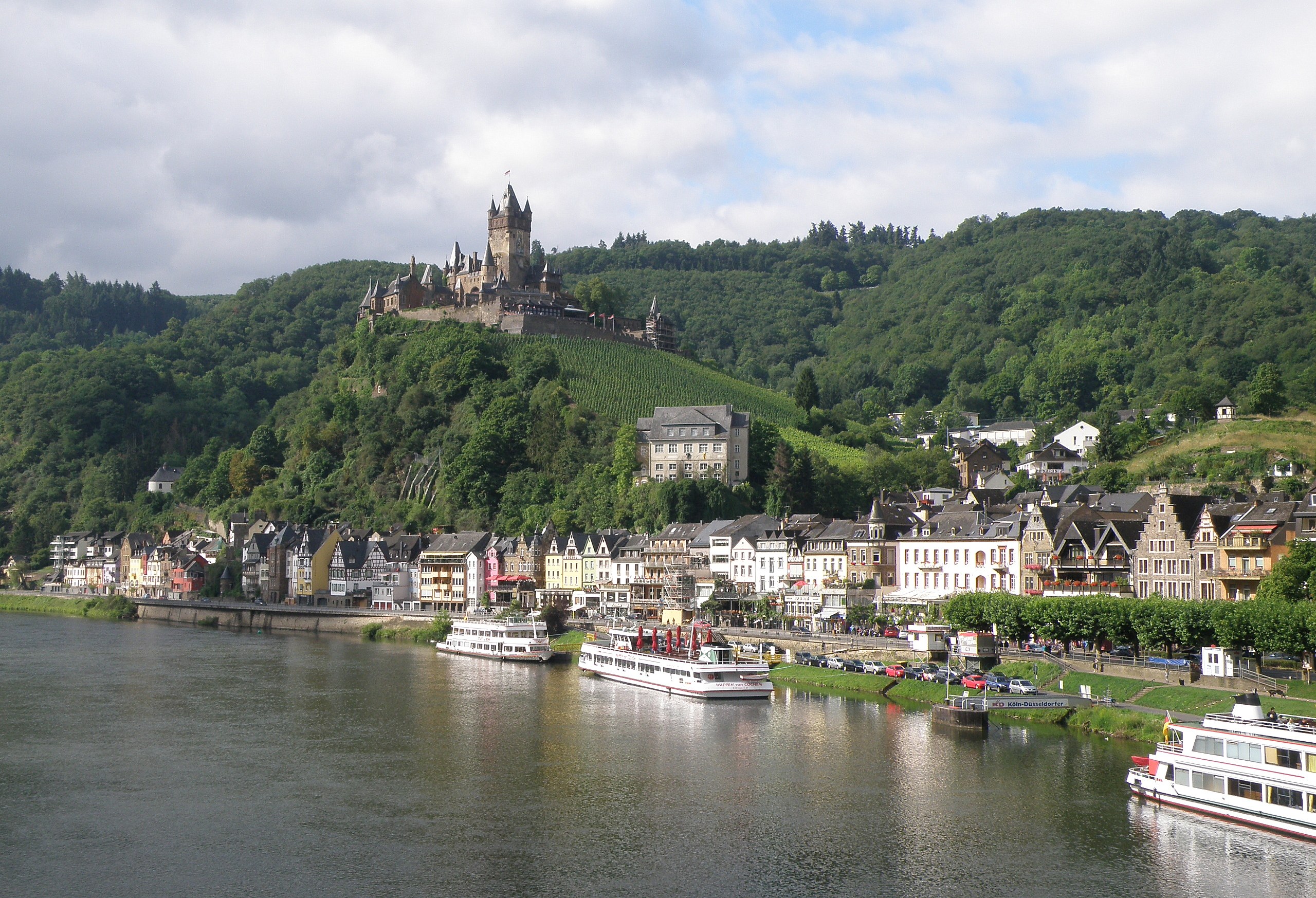 Eine malerische Aussicht auf den Rhein in Deutschland, mit einer Burg auf einem Hügel, Booten auf dem Wasser, Fahrzeugen auf einer näheren Straße und einem bewölktem Himmel.