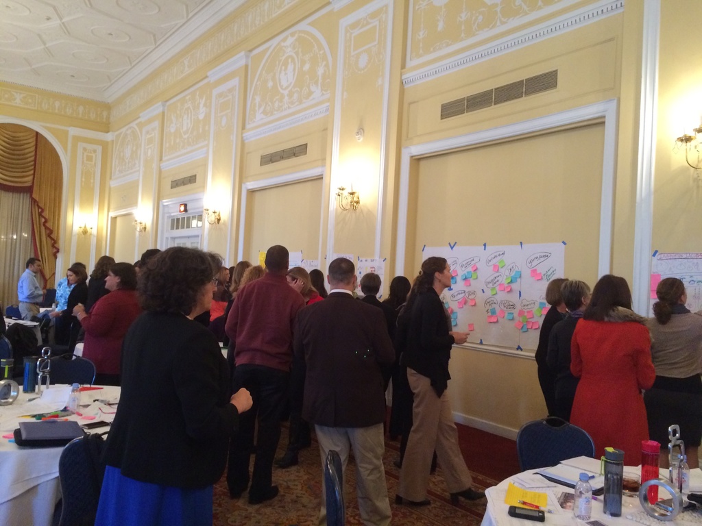 Eine Gruppe von Menschen untersucht Plakate und Diagramme, die an einer Wand in einem Raum aufgehängt zu sein scheinen, der wie ein Kunstmuseum aussieht.