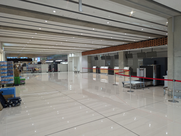 Großer Flughafen-Check-in-Bereich mit verstreuter Gepäckstücke, Gepäckwagen, Kartons, Absperrpoller mit Bändern, Säulen, wandmontierten Fernsehern und Deckenleuchten.