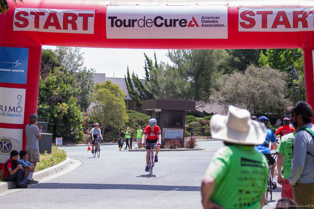 Eine belebte Straßenszene mit Menschen auf Fahrrädern, einige auf einem Gehweg sitzend und andere auf der Straße stehend, mit einer Hütte und umliegenden Pflanzen und Bäumen im Hintergrund.