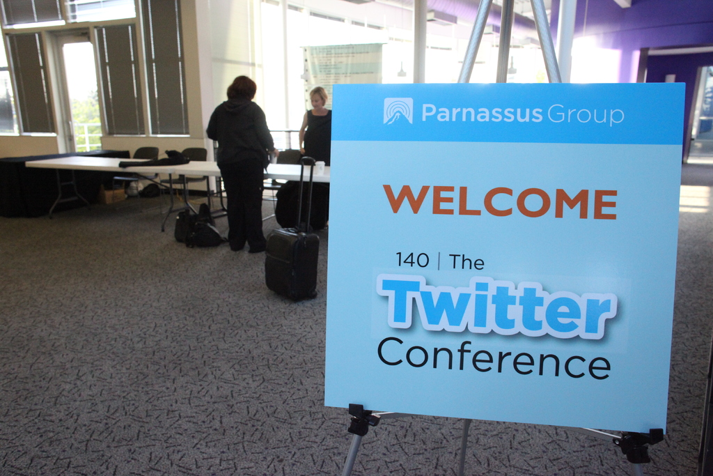 Ein Board mit "Willkommen" und "Twitter" darauf, mit ein paar Leuten, einem Tisch und mehreren Stühlen im Hintergrund.