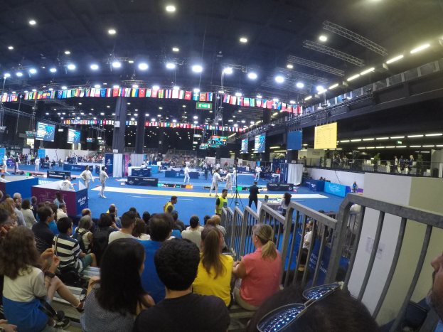 Eine große Menge sitzend und stehend in einer Indoor-Sportarena mit Bildschirmen, Fahnen und Deckenleuchten zu sehen.