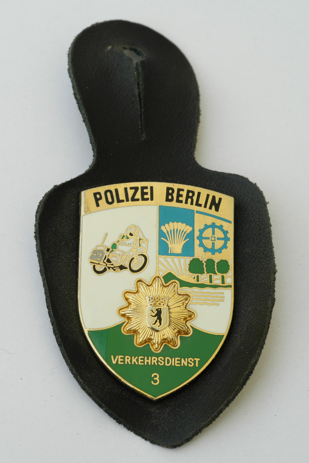 Ein schildförmiges Polizeisiegel mit 'Polizei Berlin' in schwarzer Schrift, das ein weißes Stern auf blauem Hintergrund und einen weißen Rand hat und auf einer Oberfläche liegt.