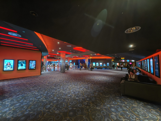 Ein großer Raum mit Menschen auf Sofas sitzend, die an der Wand montierte Fernseher beobachten, umgeben von Säulen und Deckenleuchten, mit dem Boden im Vordergrund.