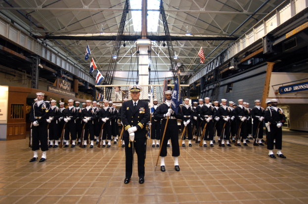 Eine Gruppe uniformierter Männer in einem großen Raum, die Mützen tragen und Gewehre halten, mit Fahnen im Hintergrund, Texttafeln an den Wänden und Deckenleuchten und Stangen.