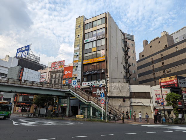 Eine belebte Stadtstraße mit einem Bus, hohen Gebäuden, Schildern, einer Brücke, Fußgängern, Fahrzeugen, einem Zaun, Bäumen, Straßenlaternen und einer bewölkten Himmel