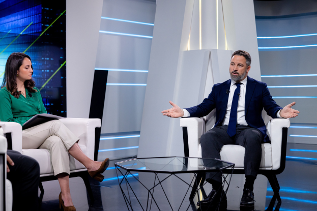 Zwei Personen sitzen auf einem TV-Sessel: ein Mann in einem blauen Blazer, weißem Hemd und blauer Krawatte und eine Frau, die ein Buch hält; ein Glas steht auf dem Tisch zwischen ihnen mit Studiobeleuchtung und einer Wand im Hintergrund.
