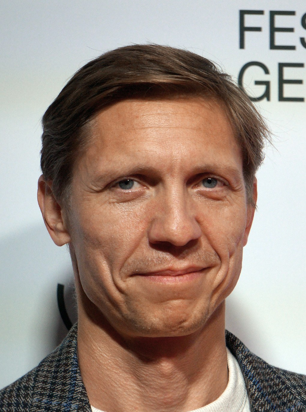 Ein Mann im Anzug lächelt in die Kamera, im Hintergrund eine Tafel mit "Eddie Redmayne Vermögen".