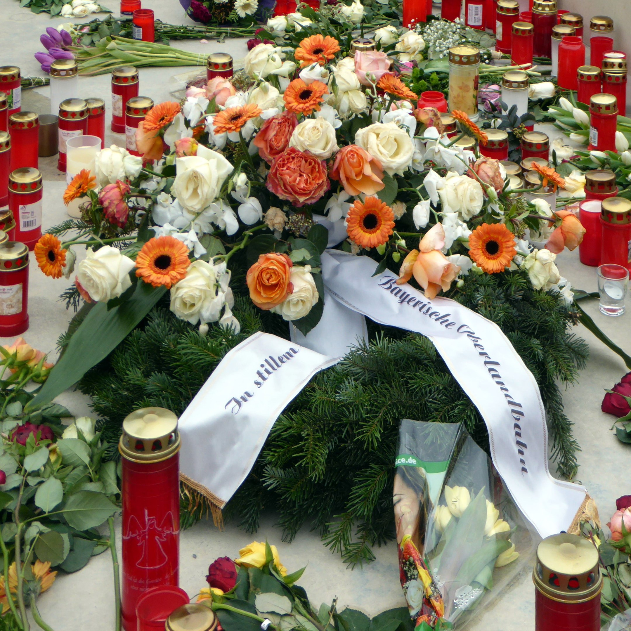 Mahnmal in Berlin mit Blumen, Kerzen und Blättern, die vor einer Wand auf dem Boden liegen.
