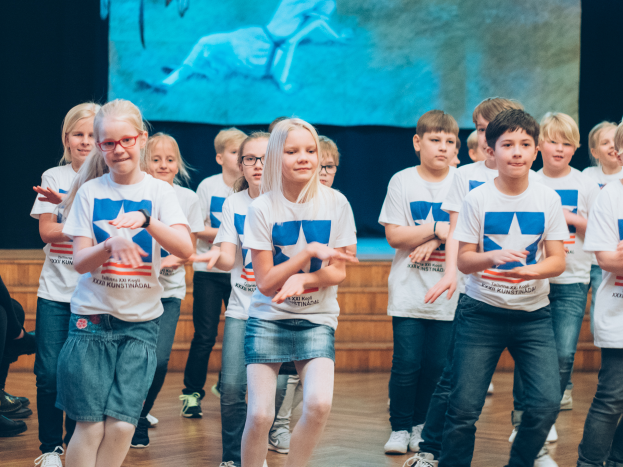 Eine Gruppe von Kindern in weißen Hemden und blauen Shorts, einige mit Brille, tanzt auf einer Bühne mit Zuschauern links und einem Bildschirm im Hintergrund, bei einem Tanzwettbewerb.